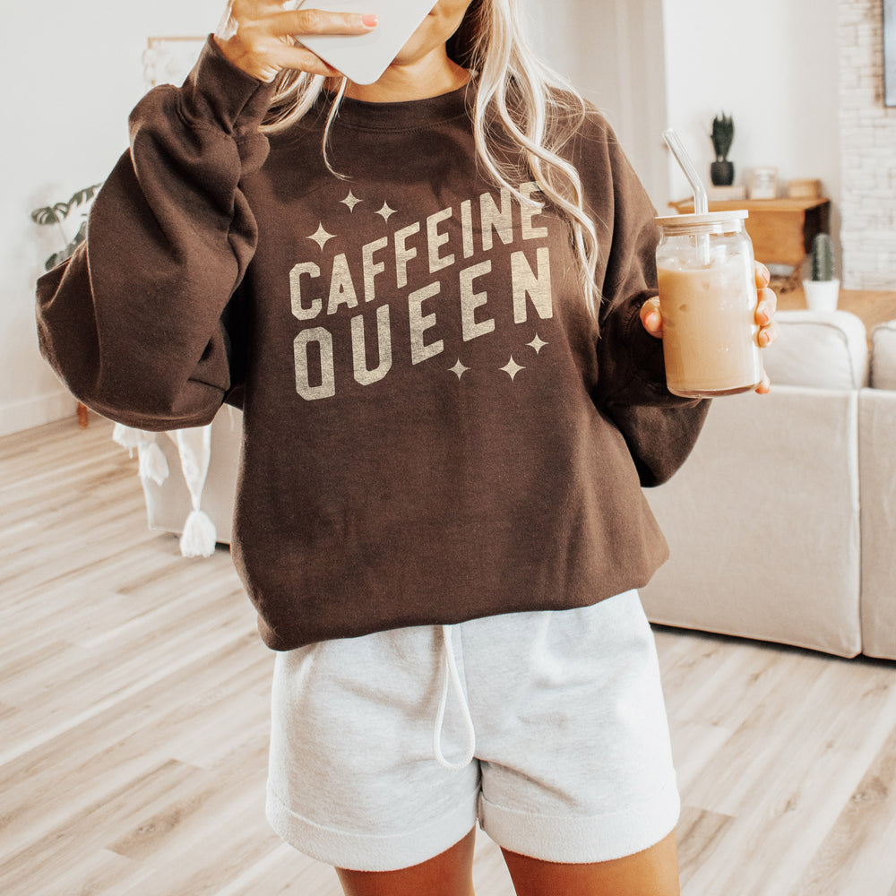 A cute young woman holding a coffee, wearing a caffeine queen sweatshirt in Dark Chocolate