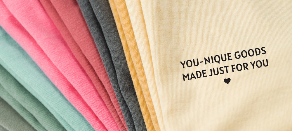 a colorful stack of t-shirts with an Yellow one on top with words that say "you-nique goods made just for you".