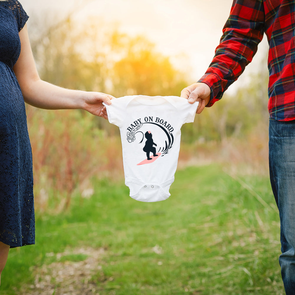 
                  
                    a couple holding a baby onesie between them, printed with Baby On Board - Surfing Girl svg as a pregnancy announcement
                  
                