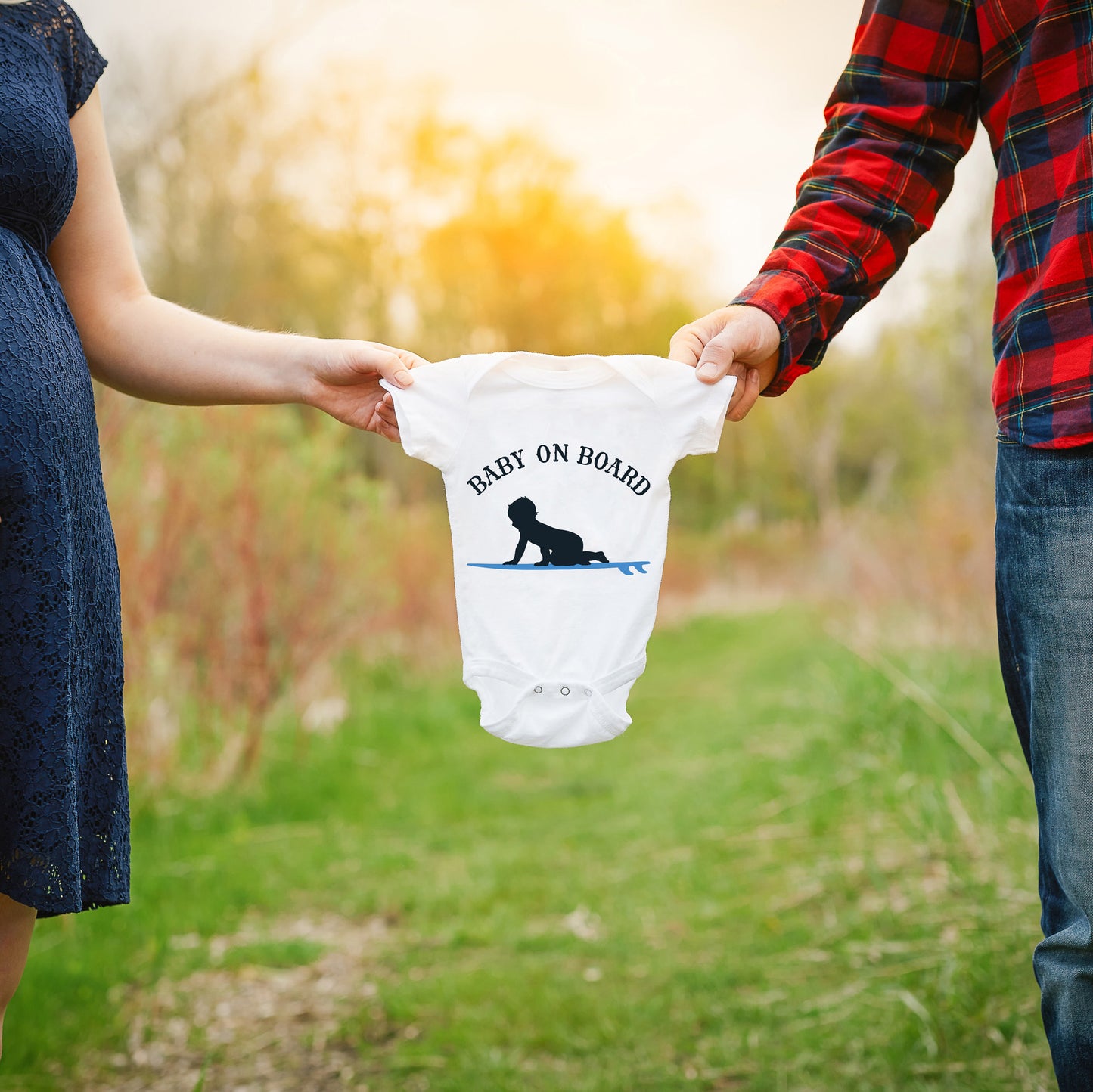 
                  
                    a couple holding a onesie between them with Baby On Board - Surf Boy svg printed in blue as a baby announcement
                  
                