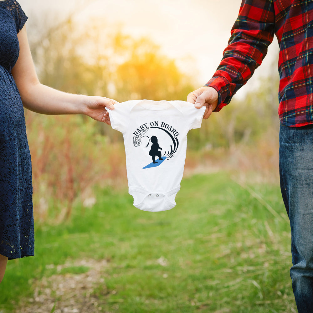 
                  
                    a couple holding a baby onesie between them, printed with the Baby On Board Surfing Boy SVG in blue, as a gender reveal announcement
                  
                