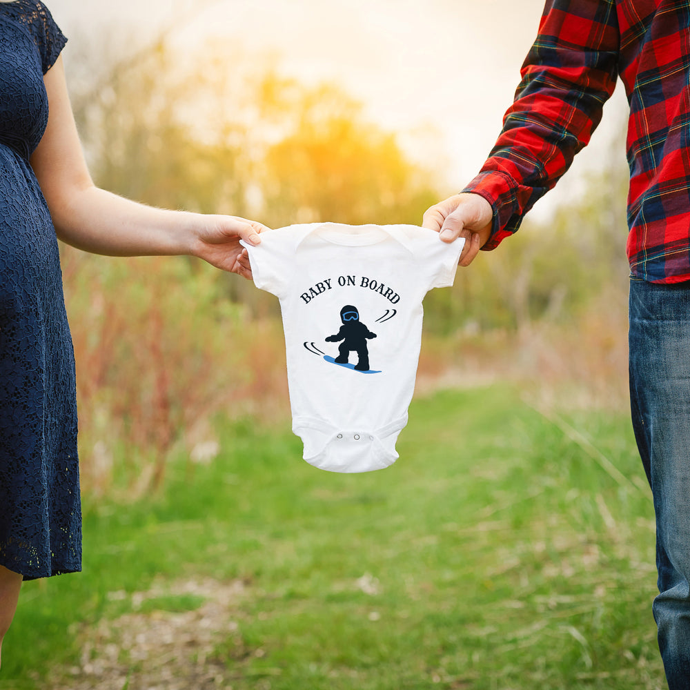 
                  
                    a couple holding a onesie between them, printed with baby on snowboard svg, as a gender reveal announcement 
                  
                