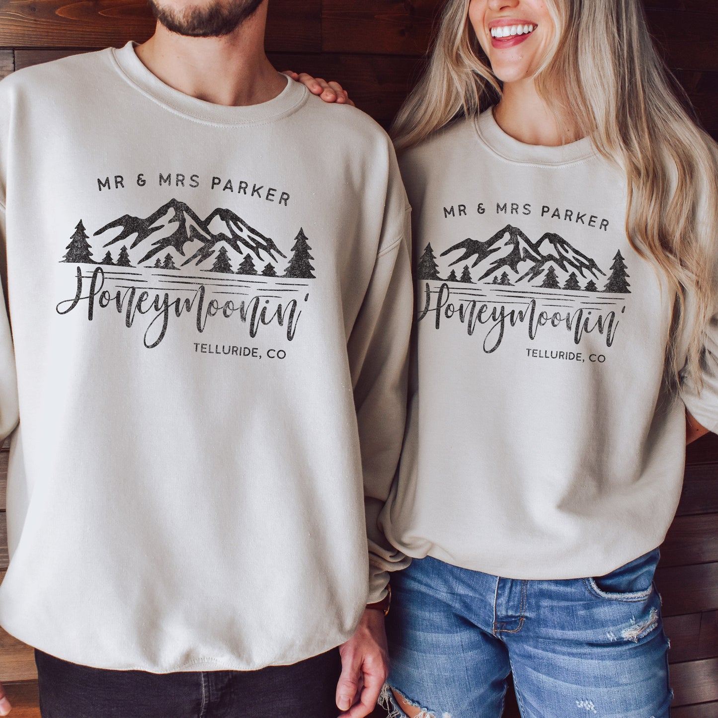 a young couple in a rustic cabin, wearing oversized honeymoonin' custom location sweatshirts in sand