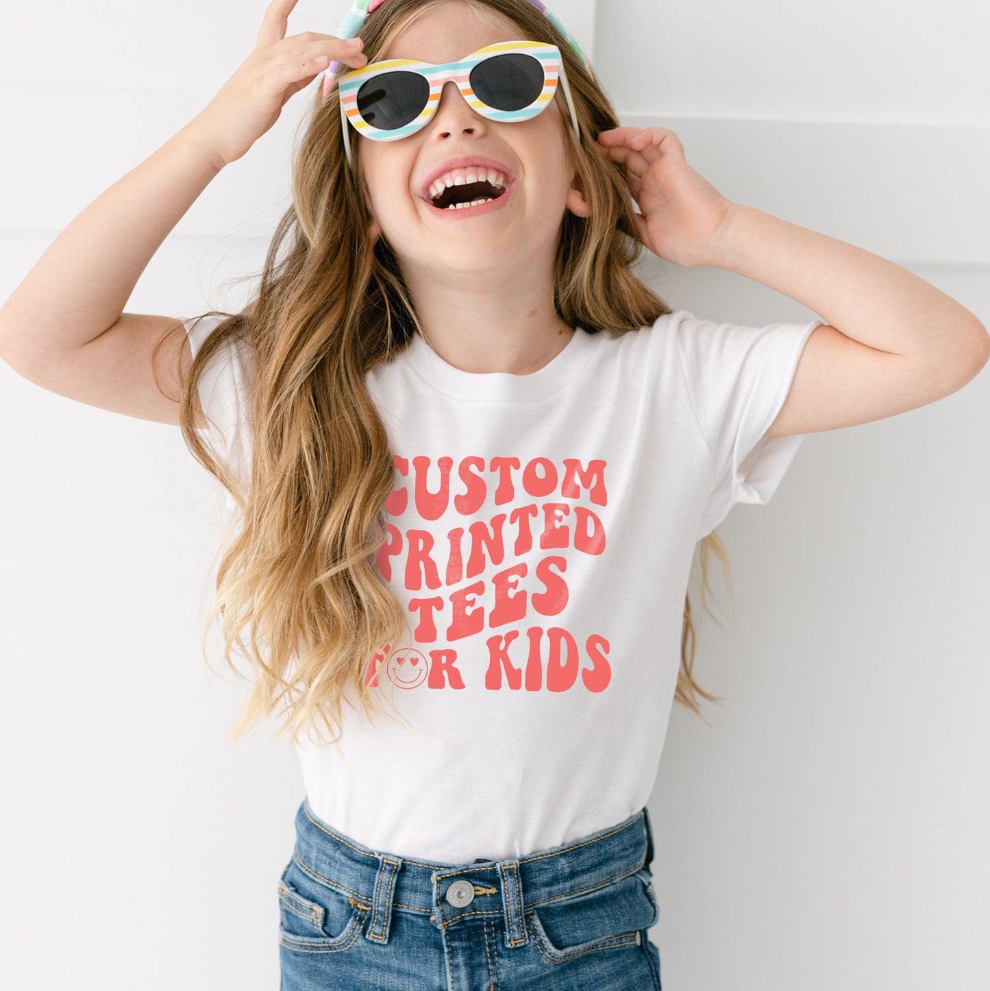 a cute smiling girl wearing sunglasses and a white t-shirt that reads "custom printed tees for kids"