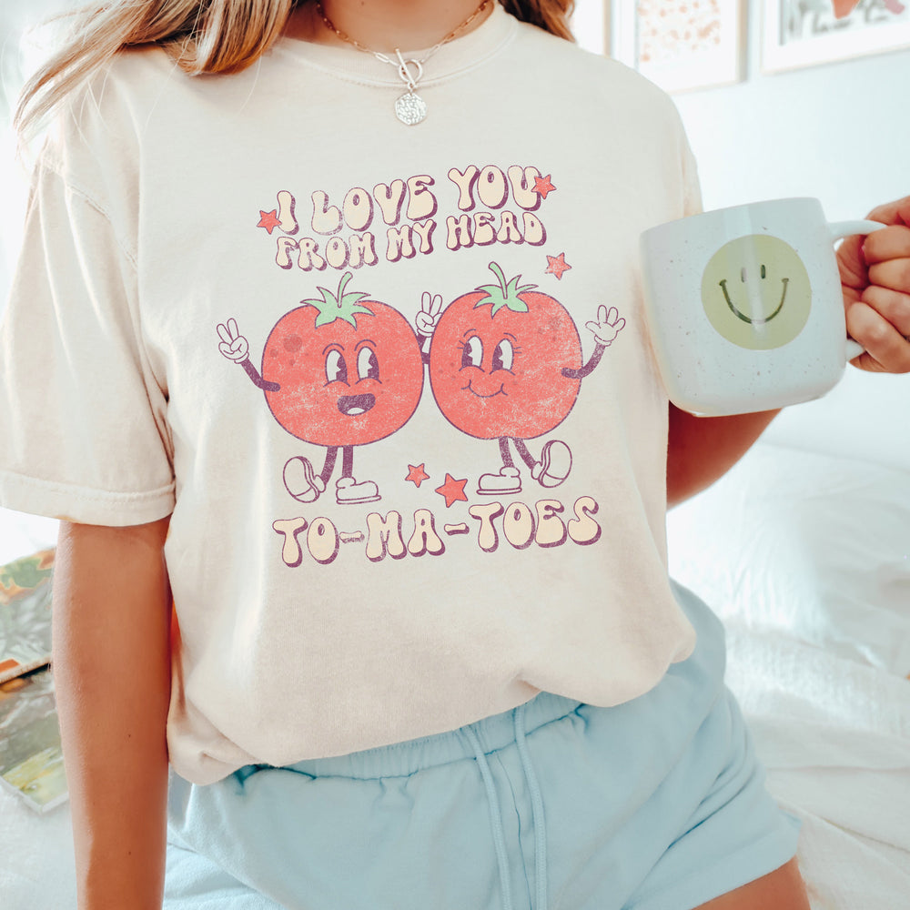 a young woman sitting on her bed, holding a smiley face coffee mug, wearing a retro I love you from my head to-ma-toes tee in Ivory