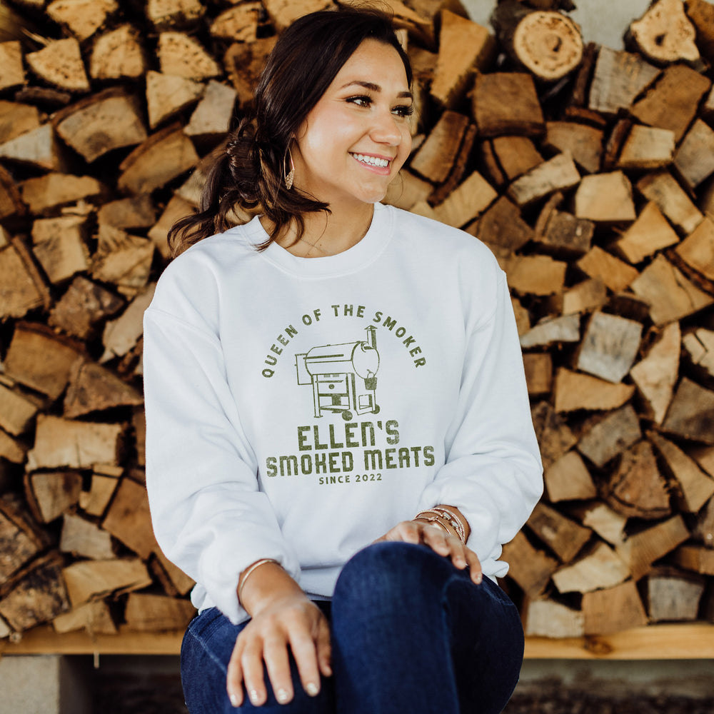 A pretty woman sitting in front of a wood stack, wearing a personalized traeger inspired sweatshirt in white