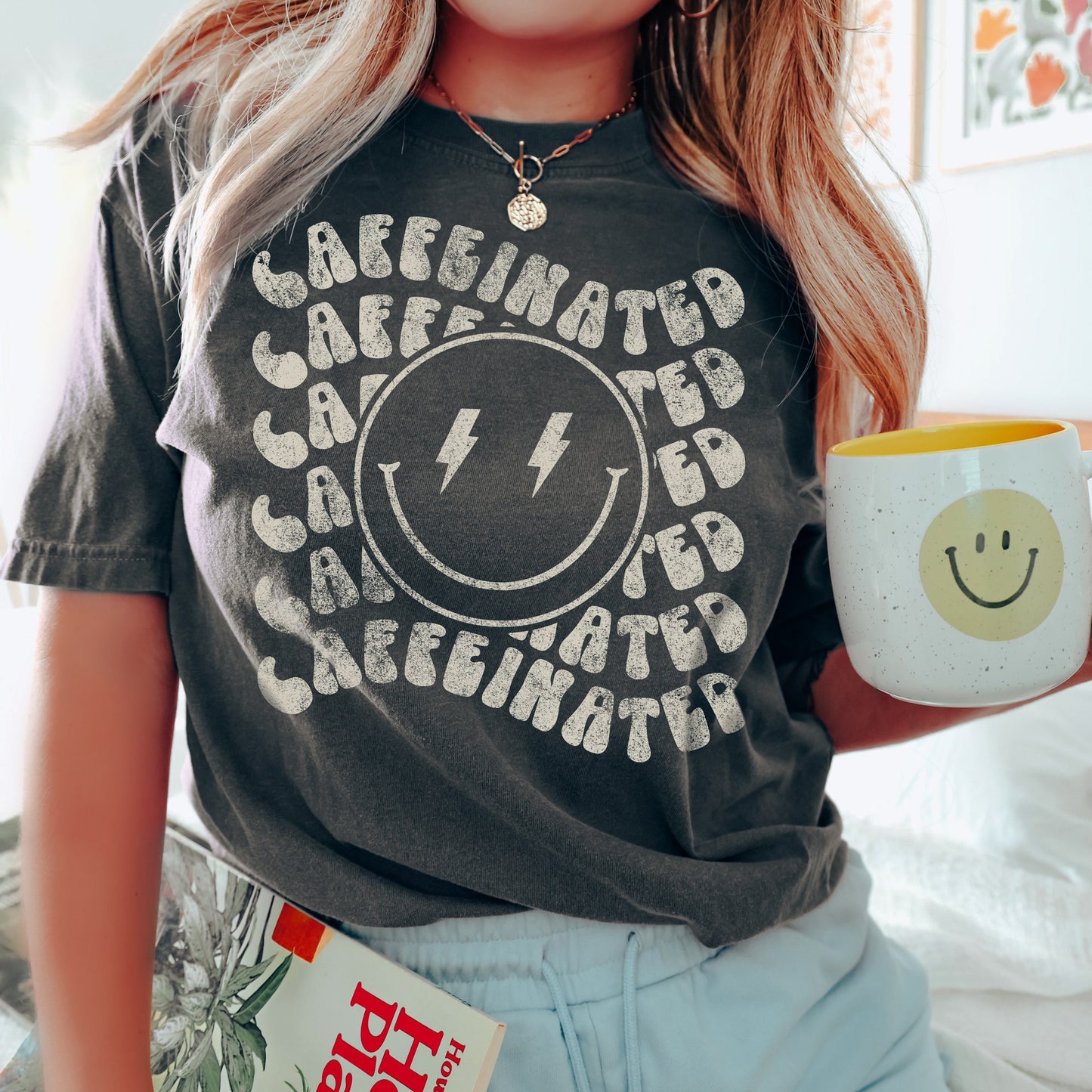 a young woman at home holding a coffee cup, wearing a caffeinated smiley tee in pepper