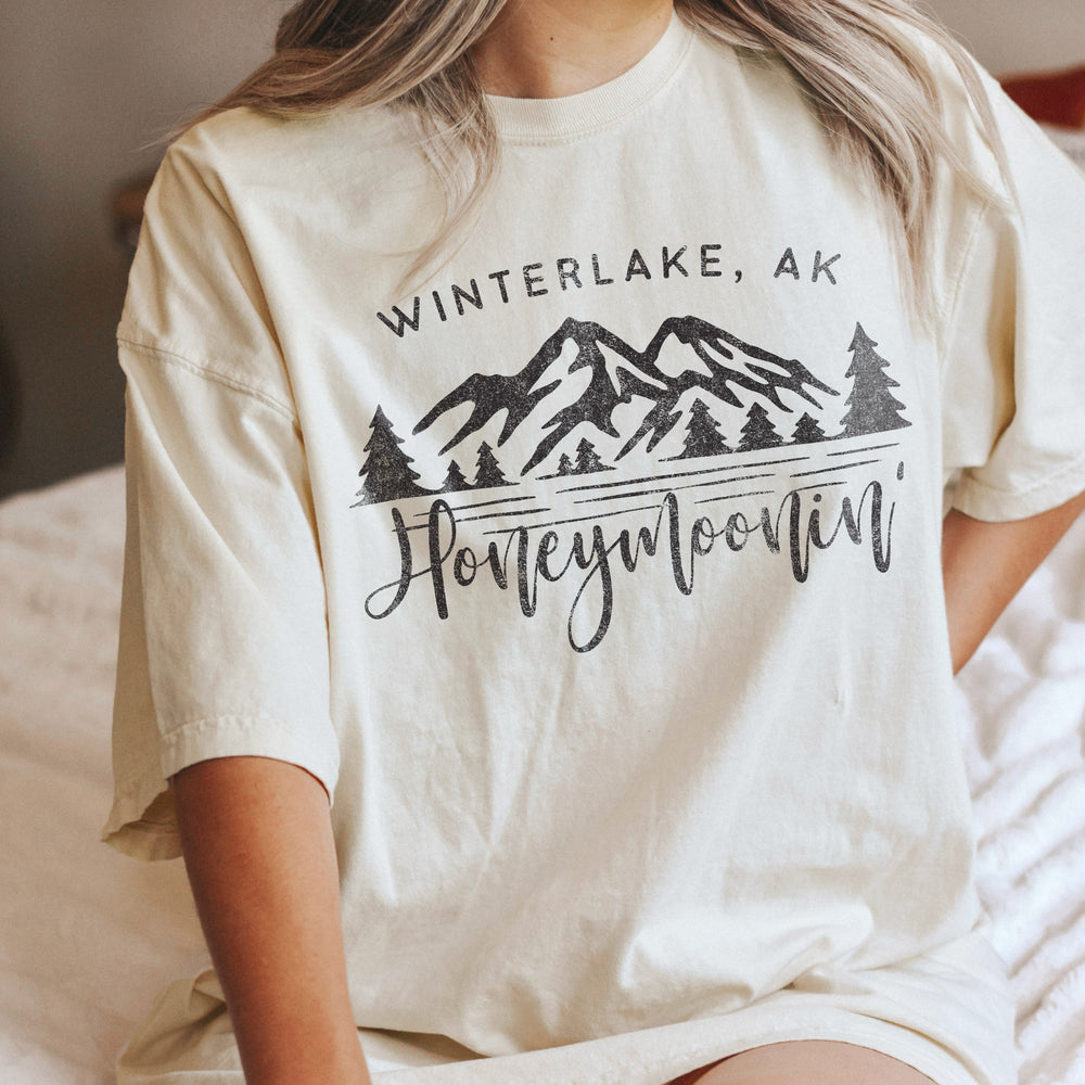a young woman in bed wearing an oversized, custom mountain location honeymoonin' tee in natural